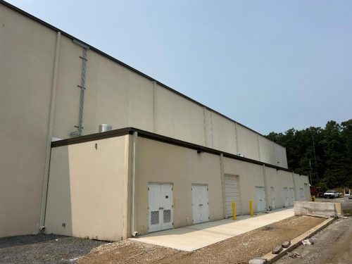 Beige industrial building with several doors, a loading dock, and a sidewalk next to a gravel area.