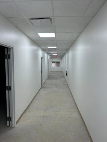 Long, empty hallway with white walls, open doors on the left, exposed concrete floor, and ceiling lights.
