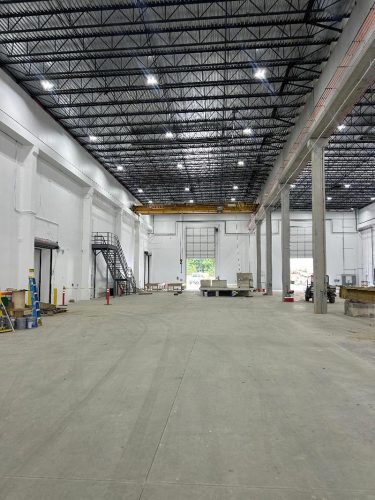 Spacious, empty warehouse with high ceilings, white walls, exposed metal beams, industrial lighting, a staircase, and large open doors at the far end. Construction materials and equipment are scattered around.