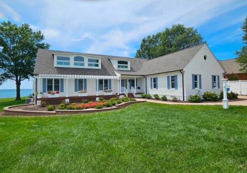 White house with large windows, manicured lawn, flower beds, and a view of water in the background.