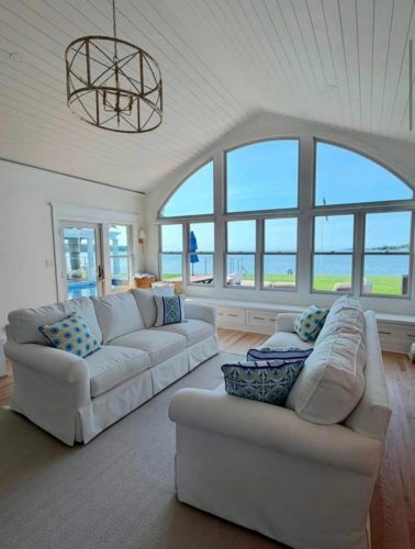 Bright living room with white sofas, patterned pillows, and large windows overlooking a lake and green lawn.