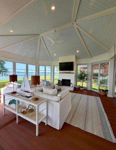 Bright sunroom with white sofas, a fireplace, TV, large windows, and a view of a lake and greenery outside.