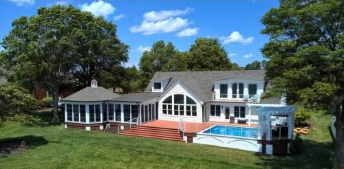 Two-story white house with a pool, large deck, and surrounded by trees and grass under a blue sky.
