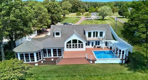 Large house with a swimming pool and deck, surrounded by trees and grassy yard, viewed from above.