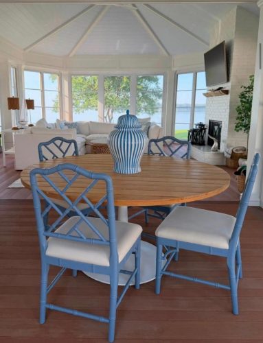 Round wooden table with blue chairs and striped vase in a bright sunroom with lake view and white furniture.