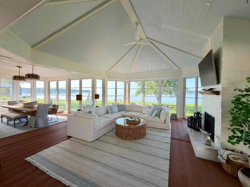 Bright sunroom with white sofas, dining table, large windows, lake view, and a fireplace with a TV above.