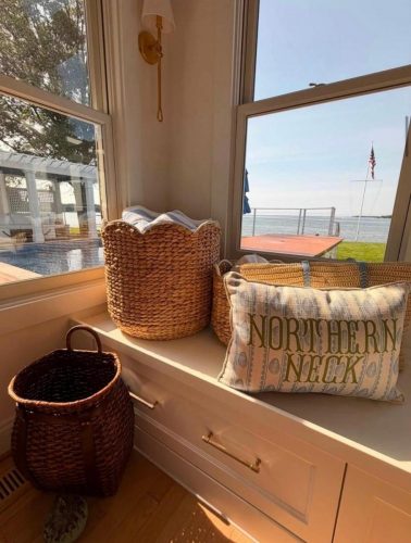 Sunlit window seat with woven baskets and a pillow that says “NORTHERN NECK,” overlooking water.