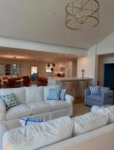 Bright open living room and kitchen with white sofas, patterned armchair, bar stools, and modern lighting.