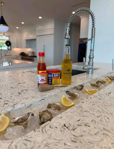 Oysters on ice with lemon wedges line a kitchen island; hot sauce, seasoning, and beer are nearby.