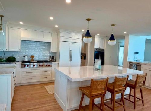 Modern white kitchen with island, pendant lights, wood floor, and three woven stools at the counter.