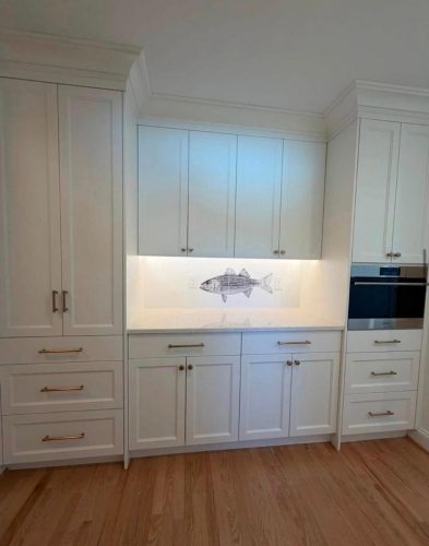 White kitchen cabinets with gold handles, wood floor, and a fish drawing on the backsplash under lighting.