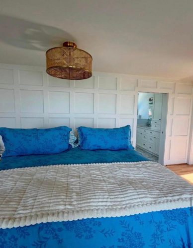 A bed with blue bedding is in front of a white paneled wall, and a doorway leads to a bathroom.