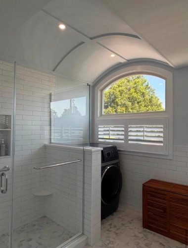 Modern bathroom with white tiles, a glass shower, washer and dryer, wooden bench, and large arched window.