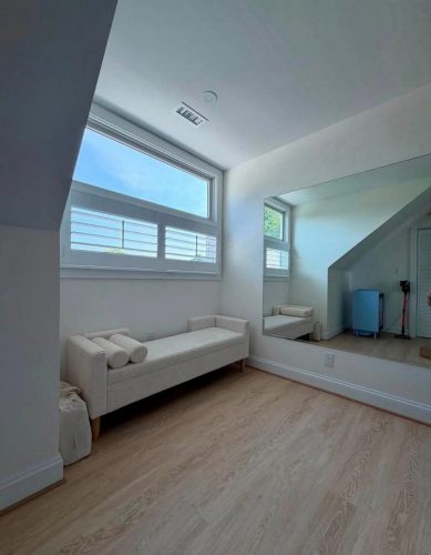 Minimalist room with a bench, mirror, wood floor, and a large window letting in daylight.