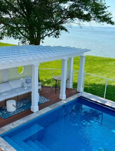 A poolside pergola with white seating overlooks a grassy lawn and a lake on a sunny day.