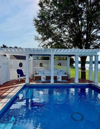 A blue pool with a white pergola, seating area, and lake view in the background.